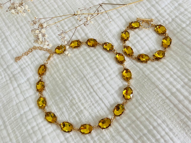 K&ouml;nnte beinhalten: Ein goldfarbenes Halsketten- und Armbandset mit ovalen, bernsteinfarbenen Steinen. Sowohl die Halskette als auch das Armband bestehen aus einer zarten goldenen Kette.