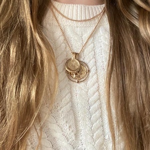 May include: A gold layered chain necklace with a pendant featuring three stacked round coins. The necklace is worn by a person with long blonde hair and a white cable knit jumper.