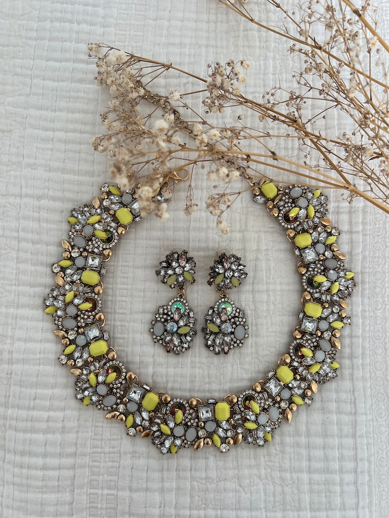 May include: A yellow and white statement necklace and matching earrings with a geometric design. The jewelry is made of metal and features a variety of clear and yellow rhinestones.