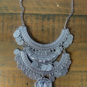 May include: A silver necklace with a large, ornate pendant featuring a clear crystal. The pendant is made up of multiple layers of silver coins and chains.