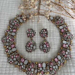 May include: A gold tone statement necklace and matching earrings with pink and white rhinestones. The necklace is a choker style and the earrings are dangle style.