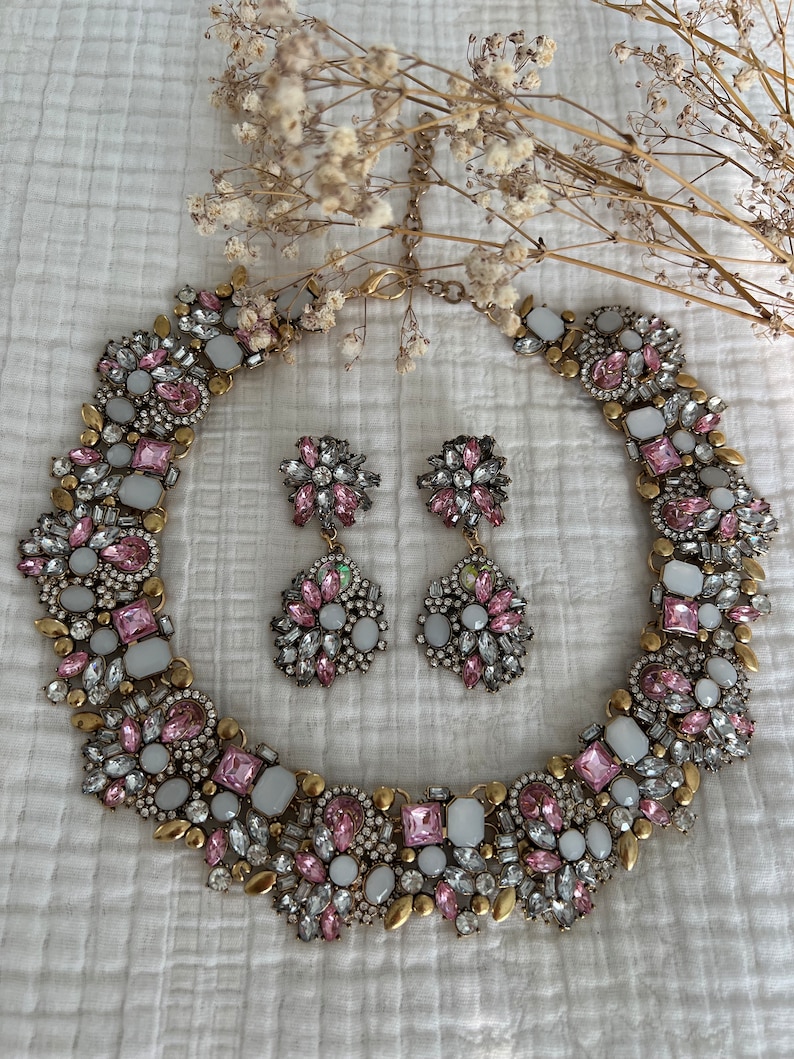 May include: A gold tone statement necklace and matching earrings with pink and white rhinestones. The necklace is a choker style and the earrings are dangle style.