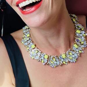 May include: A close-up of a woman wearing a gold and yellow statement necklace with a variety of clear and pastel blue stones. The necklace is made of metal and has a geometric design.