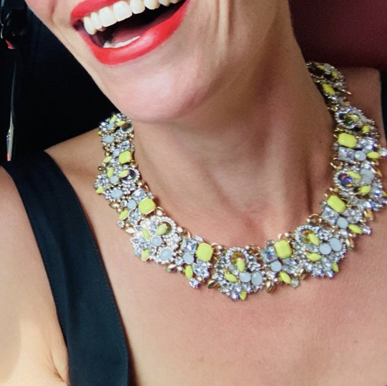 May include: A close-up of a woman wearing a gold and yellow statement necklace with a variety of clear and pastel blue stones. The necklace is made of metal and has a geometric design.