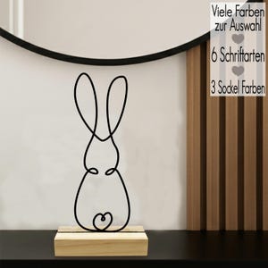 Könnte beinhalten: Eine schwarze Drahtskulptur eines Hasen, montiert auf einem kleinen Holzsockel. Der Hase ist in einer durchgehenden Linie dargestellt, mit einem herzförmigen Detail am unteren Ende. Im Hintergrund steht der Text "Viele Farben zur Auswahl", "6 Schriftarten" und "3 Sockel Farben".