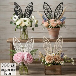 Könnte beinhalten: Blumenarrangements in Gläsern, einem Terrakottatopf und einer Holzkiste, jeweils mit dekorativen 3D-gedruckten Hasenohren verziert. Die Ohren sind in Schwarz und Weiß gehalten und weisen aufwendige florale Designs auf. Die Blumen sind in verschiedenen Farben.