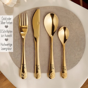 Könnte beinhalten: Gold- oder Silber-Kinderbesteck mit Messer, Gabel und zwei Löffeln. Das Besteck ist mit dem Namen "Victoria" und einem kleinen Tiermotiv graviert. Der Text "Gold oder Silber Farben", "8 Schriftarten zur Auswahl", und "Hochwertige Lasergravur" ist auf dem Bild zu sehen.