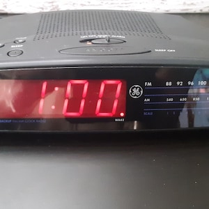 Puede incluir: Un radio despertador digital negro con una pantalla digital roja que muestra la hora 1:00. El radio despertador tiene una función de respaldo de batería e incluye bandas de radio AM y FM.