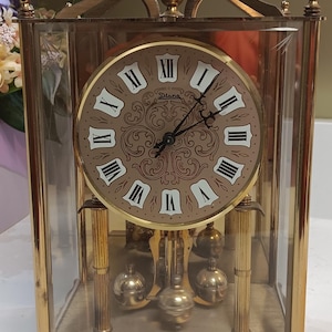 May include: A gold-coloured, rectangular clock with a glass enclosure. The clock face is tan with black Roman numerals and the brand name "Diana". The pendulum features three gold-coloured spheres.
