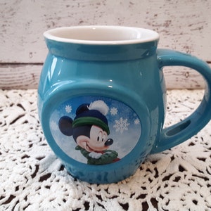 Pu&ograve; includere: Una tazza in ceramica turchese con Topolino con un cappello e una sciarpa invernali. La tazza ha una forma arrotondata con un manico grande e un interno bianco. I fiocchi di neve sono sullo sfondo dell'immagine di Topolino.