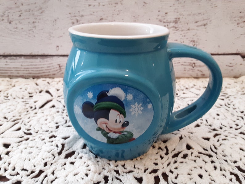 Pu&ograve; includere: Una tazza in ceramica turchese con Topolino con un cappello e una sciarpa invernali. La tazza ha una forma arrotondata con un manico grande e un interno bianco. I fiocchi di neve sono sullo sfondo dell'immagine di Topolino.