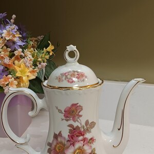 May include: White porcelain coffee pot with a lid, adorned with pink and yellow floral designs and gold trim. The handle and spout are elegantly curved. A decorative piece for serving coffee or tea.