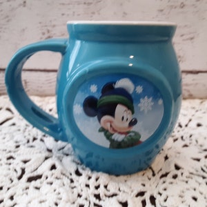 Pu&ograve; includere: Una tazza in ceramica turchese con interno bianco e manico grande. La tazza presenta un'immagine ovale di Topolino con cappello e sciarpa invernali, con fiocchi di neve sullo sfondo. La tazza &egrave; appoggiata su una superficie di pizzo bianco.