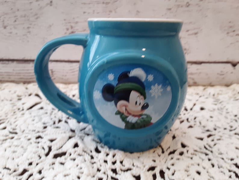 Pu&ograve; includere: Una tazza in ceramica turchese con interno bianco e manico grande. La tazza presenta un'immagine ovale di Topolino con cappello e sciarpa invernali, con fiocchi di neve sullo sfondo. La tazza &egrave; appoggiata su una superficie di pizzo bianco.