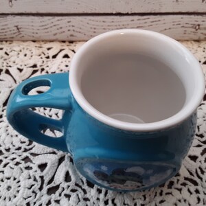 Pu&ograve; includere: Una tazza in ceramica turchese con interno bianco. La tazza ha un design unico del manico con un foro e una piccola immagine sul lato. La tazza &egrave; appoggiata su un centrino di pizzo bianco.