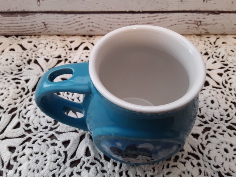 Pu&ograve; includere: Una tazza in ceramica turchese con interno bianco. La tazza ha un design unico del manico con un foro e una piccola immagine sul lato. La tazza &egrave; appoggiata su un centrino di pizzo bianco.