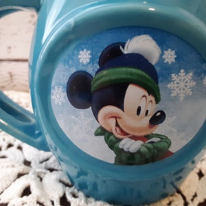 Pu&ograve; includere: Una tazza in ceramica azzurra con un design di Topolino a tema invernale. Mickey indossa un cappello blu con un pompon bianco e una sciarpa verde. La tazza ha un manico grande e un design rotondo.
