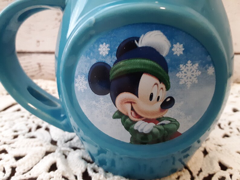 Pu&ograve; includere: Una tazza in ceramica azzurra con un design di Topolino a tema invernale. Mickey indossa un cappello blu con un pompon bianco e una sciarpa verde. La tazza ha un manico grande e un design rotondo.