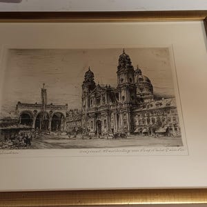 May include: A framed black and white etching depicting a detailed architectural scene of a large cathedral with a prominent dome and towers, set in a public square with arches. The artwork is enclosed in a metallic gold frame with a cream mat.