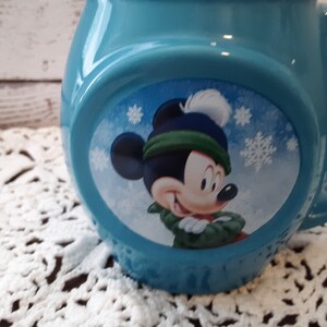 Pu&ograve; includere: Una tazza in ceramica azzurra con un design invernale di Topolino. Topolino indossa un cappello e una sciarpa verdi e blu, con fiocchi di neve. La tazza ha una forma arrotondata e un manico grande.