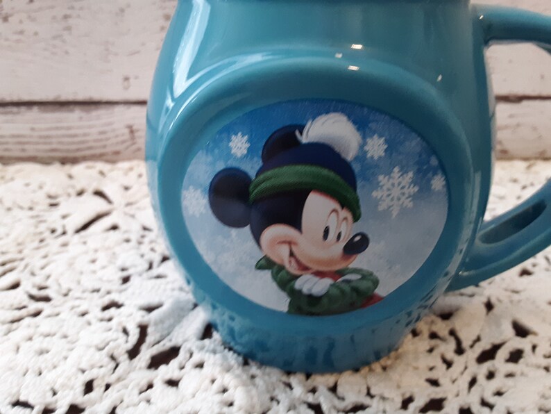 Pu&ograve; includere: Una tazza in ceramica azzurra con un design invernale di Topolino. Topolino indossa un cappello e una sciarpa verdi e blu, con fiocchi di neve. La tazza ha una forma arrotondata e un manico grande.