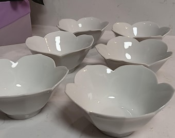 May include: Six white ceramic bowls with a petal-like design. The bowls are arranged on a white surface. The bowls have a glossy finish and are empty.