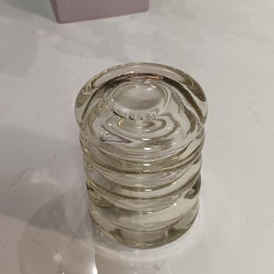 May include: Clear glass object with a cylindrical shape and multiple horizontal ridges. The object is sitting on a white surface. In the background, a purple and gray vase holds flowers.