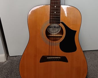 May include: A light brown acoustic guitar with a dark brown fretboard and a black teardrop-shaped pickguard. The sound hole features a circular rosette pattern and a white label inside with the text Overload Musical Instruments printed clearly.