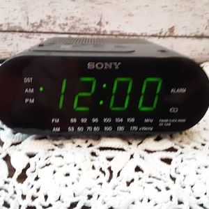 Op de afbeelding: Zwarte digitale Sony-wekker met groene cijfers 12:00. De klok heeft FM- en AM-radiofrequentie-indicatoren. Het woord "SONY" staat op de bovenkant van de klok.