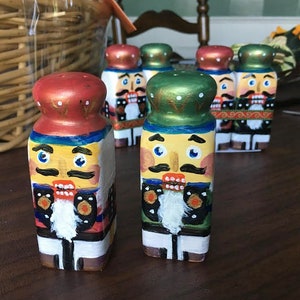 May include: A set of six hand-painted nutcracker salt and pepper shakers. The shakers are made of wood and are painted in a variety of colors, including red, green, blue, yellow, and white. Each shaker has a different facial expression and a different hat.