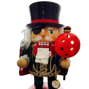 May include: A decorative wooden nutcracker figurine. It features a black top hat with a red band, an eye patch, and a white mustache. The figurine wears a green and red coat with gold accents and holds a red ball ornament. It stands on a red base.