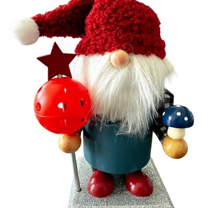 May include: A whimsical gnome figurine with a red knit hat and beard. The gnome holds a red star-topped staff and a blue mushroom. The figurine is wearing a teal tunic and red boots, standing on a glittery base. A festive holiday decoration.