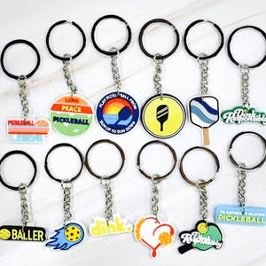 May include: A set of 12 colorful pickleball keychain charms with different designs, including a paddle, a ball, and phrases like "Peace Pickleball" and "Drink Responsibly".