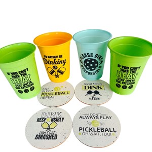 May include: A collection of four plastic cups in green, yellow, and blue, each with a different pickleball-themed phrase. Below the cups are four matching coasters with pickleball-related sayings. The text includes phrases like "Dink Responsibly" and "I'd Rather Be Dinking."