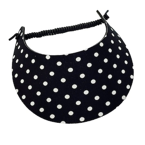May include: A black and white polka dot visor with a black elastic band. The visor is made of fabric and has a curved brim. The white polka dots are evenly spaced across the black background. The visor is designed to be worn on the head.