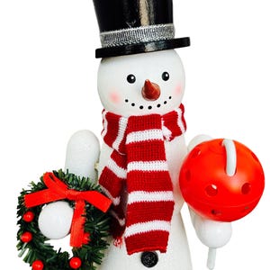 May include: A snowman figurine with a black top hat, a red and white striped scarf, and a carrot nose. The snowman holds a small green wreath with a red bow and a red ball with a white handle. The snowman has black button eyes and a coal smile.