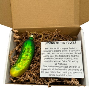 May include: A green and yellow pickle-shaped Christmas ornament inside a cardboard box with shredded paper. The ornament has a gold string for hanging. A card reads "LEGEND OF THE PICKLE" and describes the tradition of the pickle ornament.