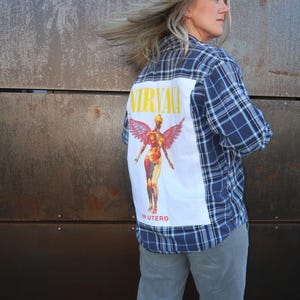 Könnte beinhalten: Blau kariertes Flanellhemd mit einem weißen Rückenteil mit dem Nirvana "In Utero" Albumcover. Die Grafik zeigt den Bandnamen in Gelb und eine stilisierte anatomische Figur mit Flügeln.
