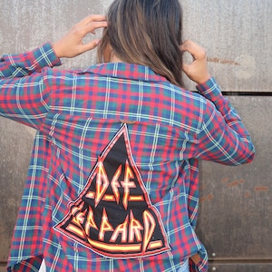 Peut inclure: Chemise à carreaux rouge, bleu et vert avec un patch noir au dos portant le logo Def Leppard en jaune et orange. Portée avec un jean en denim bleu.
