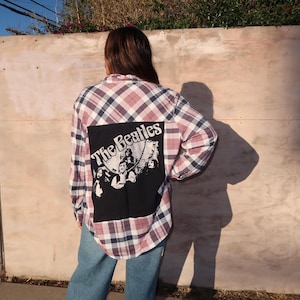 May include: A plaid button-down shirt in shades of pink, white, and navy blue. The back of the shirt features a black patch with a graphic of The Beatles and their band name in white. The shirt is worn with blue jeans.