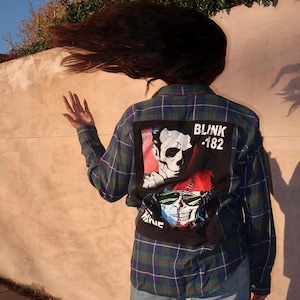 May include: A plaid flannel shirt in shades of blue, green, and red, featuring a graphic patch on the back with the text "BLINK -182" and a skull design. The shirt is worn with blue jeans.
