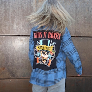 Może przedstawiać: Niebieska koszula w kratę z flaneli z dużą czarną naszywką z logo Guns N' Roses i grafiką czaszki. Tekst "GUNS N' ROSES" jest czerwony. Koszula ma długie rękawy i klasyczny kołnierz.