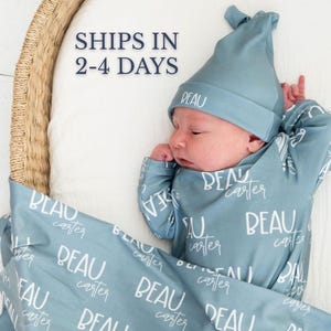 May include: A newborn baby wearing a light blue hat, sleeper, and blanket. The blanket and sleeper are patterned with the name "BEAU" and "carter" in white script. The hat has "BEAU" embroidered on it. The baby is lying in a wicker basket.