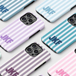 Puede incluir: Una colección de fundas para teléfonos con un patrón de rayas en varios colores, incluyendo rosa, azul y morado. Cada funda presenta las iniciales "JKF" en un color a juego. Las fundas están diseñadas para un smartphone.