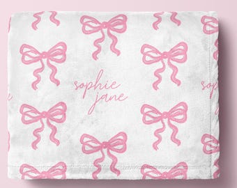 May include: A white plush blanket featuring a repeating pattern of pink hand-drawn style bows. The center of the blanket displays the name Sophie Jane written in a cursive pink script font. The fabric has a soft, textured appearance throughout.