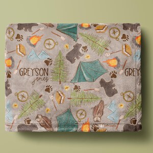 May include: A gray blanket with a camping-themed pattern. The design includes bears, tents, compasses, campfires, trees, and paw prints. The blanket has the names "Greyson" and "Grey" printed on it.