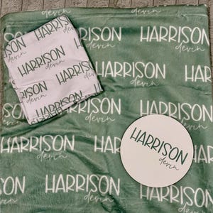 K&ouml;nnte beinhalten: Eine salbeigr&uuml;ne Pl&uuml;schdecke mit einem sich wiederholenden wei&szlig;en Muster des Namens Harrison Devin, ein gefaltetes wei&szlig;es Stoffquadrat mit demselben gr&uuml;nen Text und eine runde wei&szlig;e Holzscheibe, die mit dem Namen Harrison Devin in Gr&uuml;n bedruckt ist.