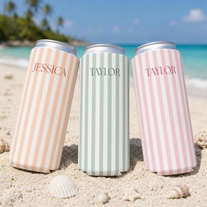 May include: Three can coolers with vertical stripes in peach, mint green, and pink, each with a name printed on them. The peach cooler says "Jessica", while the other two say "Taylor". The coolers are on a sandy beach with seashells.