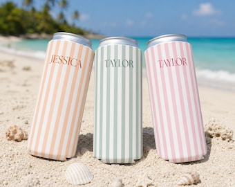 May include: Three can coolers with vertical stripes in peach, mint green, and pink, each with a name printed on them. The peach cooler says "Jessica", while the other two say "Taylor". The coolers are on a sandy beach with seashells.