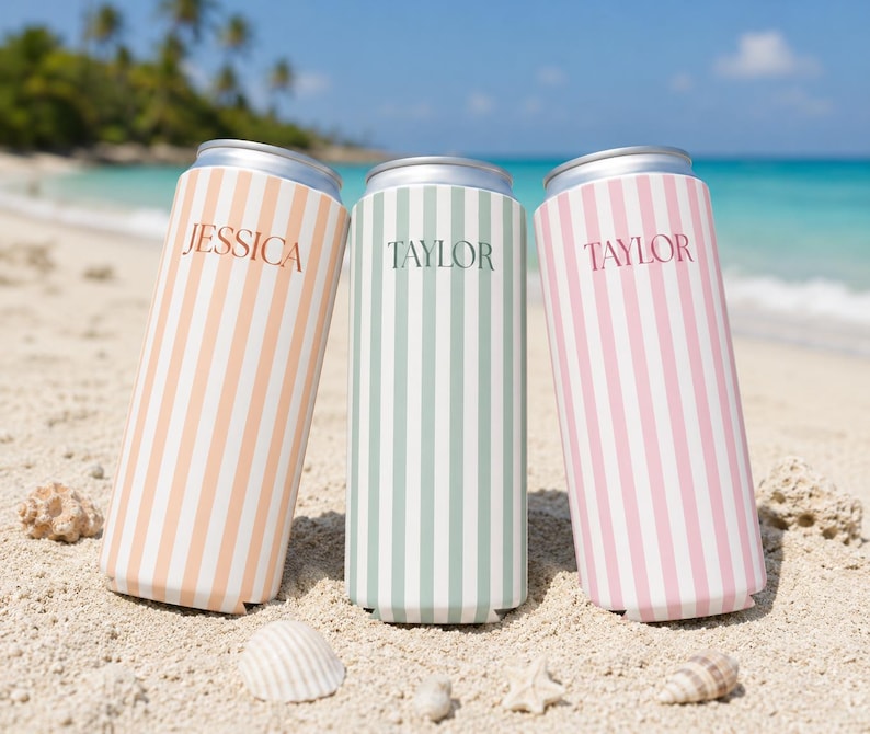 May include: Three can coolers with vertical stripes in peach, mint green, and pink, each with a name printed on them. The peach cooler says "Jessica", while the other two say "Taylor". The coolers are on a sandy beach with seashells.
